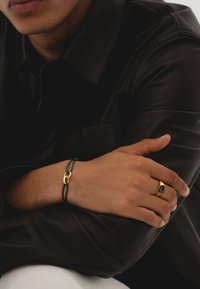 Bracelet en or avec un cordon vert, doté d'un fermoir en or. Une bague en or avec un design ovale à la main opposée. Chemise en cuir foncé.