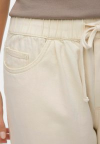 Beige bikses ar elastīgu jostu un auklu, ar priekšējo kabatu un teksturētu audumu. Tuvplāns uz jostas un kabatu.