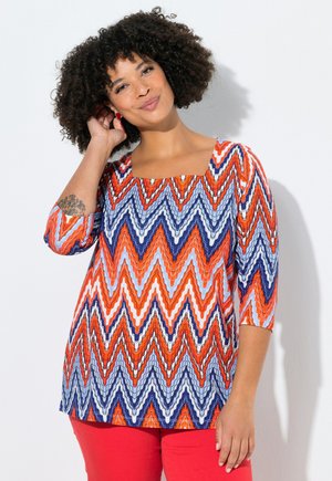 Nainen kiharat hiukset, hänellä on värikäs sahalaitakuosinen paita punaisena, sinisenä ja valkoisena, punaiset housut ja punaiset korvakorut, hymyilee ja koskettaa hiuksiaan.