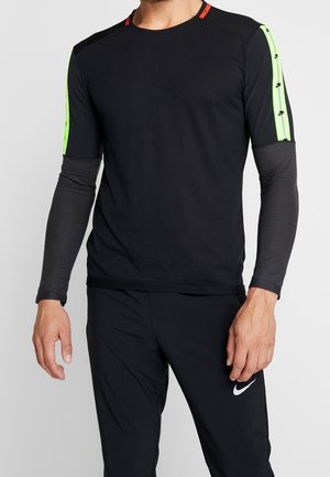 Homme portant une chemise noire à manches longues superposée avec des rayures vert néon et le logo Nike sur les manches, associée à un pantalon noir avec le logo Nike blanc.