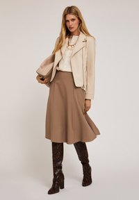 Giacca da motociclista in pelle beige sopra una blusa avorio, abbinata a una gonna A-line marrone e stivali al ginocchio marroni con stampa pitone. Accento con collana dorata.