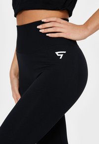 Čierne legíny s vysokým pásom, vyrobené z elastického materiálu. Obsahujú biele logo na ľavom boku a majú hladkú textúru.