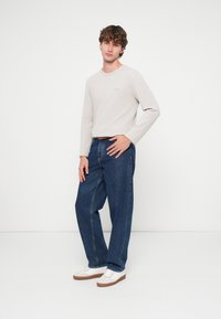 Μακρυμάνικο πουκάμισο σε ανοιχτό μπεζ χρώμα με διακριτικό λογότυπο, σε συνδυασμό με άνετο μπλε τζιν παντελόνι. Τα παπούτσια είναι λευκά αθλητικά με καφέ σόλες.