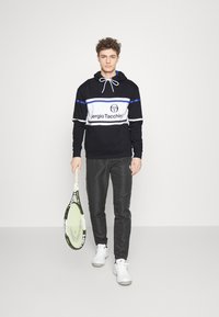 Černobílá mikina s modrými pruhy, se znakem. V kombinaci s černými kalhotami a bílými teniskami, drží tenisovou raketu.