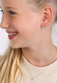 Silberne Creolen mit kleinen herzförmigen Anhängern. Das Mädchen hat lange blonde Haare und trägt einen hellen Pullover. Die natürliche Hautstruktur ist sichtbar.