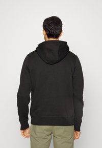 Sudadera negra con capucha ajustable, textura suave y mangas largas; combinada con pantalones de un verde oliva claro, mostrada desde atrás.