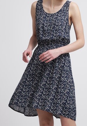 Femme portant une robe sans manches bleu marine avec un petit motif floral blanc et rose, une taille élastique et un ourlet asymétrique.