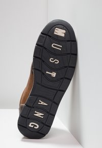 Semelle de botte marron avec du caoutchouc noir texturé, présentant des motifs moulés et le mot "MUSTANG" en lettres métalliques sur la zone du talon.