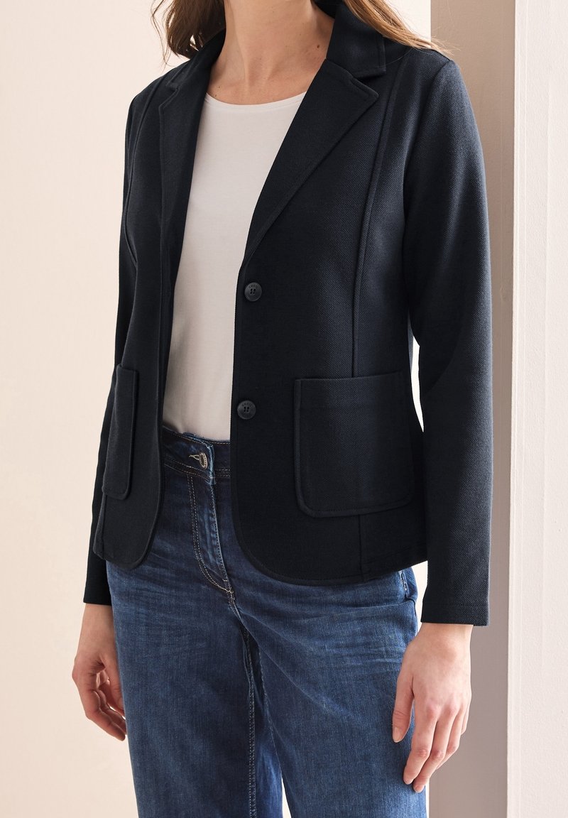 Námořnický blejzr vyrobený z texturované tkaniny s vykrojeným límcem, dvěma předními kapsami a zapínáním na knoflíky, zkombinovaný s bílým topem a džíny.