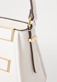 Weiße Lederhandtasche mit strukturiertem Finish, dunkelbraunen Akzenten, goldfarbener Hardware und einer strukturierten Form mit rechteckigen Details.