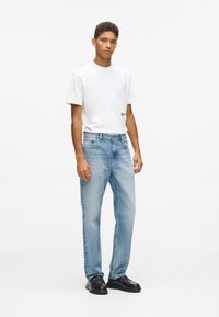 Bjela pamučna majica s kratkim rukavima i logom, uparen s svijetloplavim trapericama. Crne kožne cipele. Jednobojna bijela pozadina.