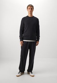 Man standing in relaxed black ribbed sweater, loose black pants, white sneakers with black stripes, and a white undershirt visible at hem.