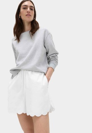 Kvinde iført grå langærmet sweatshirt og hvide shorts med bølget kant og hænderne i lommerne, står mod en ensfarvet baggrund.