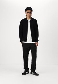 Svart zip-up jakke over en hvit t-skjorte, svarte bukser og svarte joggesko med hvite striper, accessorert med rund solbriller.