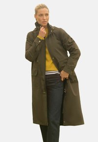Voděodolný dlouhý kabát v olivově zelené barvě, s vysokým límcem, stahovací šňůrkou, bočními kapsami a zipem; v kombinaci se žlutým svetrem a modrými džíny.