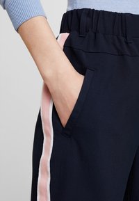 Pantalones deportivos azul marino con una franja lateral rosa y blanca, que incluyen un bolsillo y una cintura elástica. Tela suave con textura lisa.