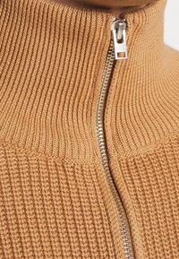 Beżowy dzianinowy sweter zapinany na suwak z wysokim kołnierzem, prążkowaną teksturą i metalicznym suwakiem z przodu.