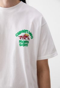 T-shirt bianca in cotone con un logo grafico verde e rosso raffigurante un personaggio in movimento, con testo che recita "Carhartt WIP" e "Pronto Subito."