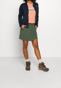 Persona vistiendo una chaqueta con cremallera azul marino, una camiseta de Patagonia color durazno, una falda verde olivo con cordón, calcetas beige y botas de senderismo marrones.