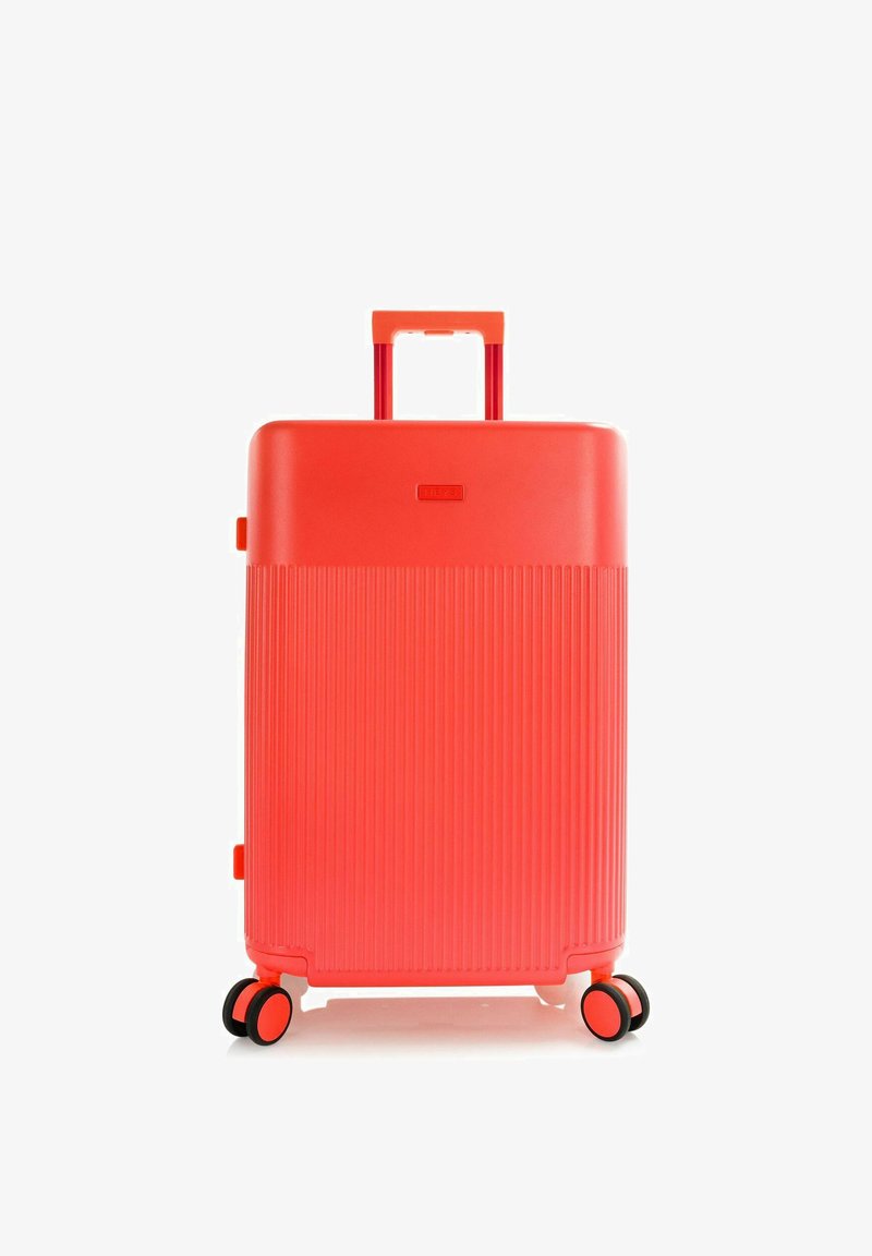 Valise rigide rouge vif avec poignée télescopique et quatre roues doubles pivotantes, debout sur fond blanc.