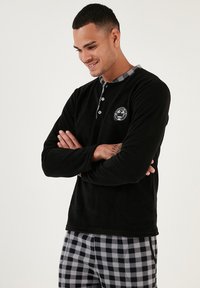 Camisa de felpa negra de manga larga con cuello abotonado y logotipo, combinada con pantalones de pijama a cuadros en gris y negro. Textura suave, ajuste relajado.