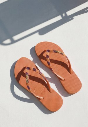 Paire de tongs orange Havaianas avec des brides à motifs projetant des ombres sur une surface blanche sous une lumière vive.