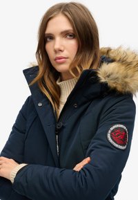 Chaqueta tipo puffer navy con capucha con pelo, que presenta un cierre de cremallera, tejido texturizado y un parche de logo rojo en la manga.