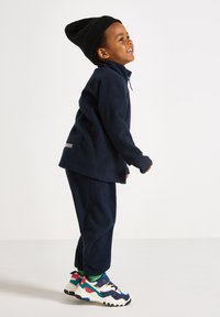 Giacca e pantaloni in fleece blu navy con colletto alto, abbinati a un berretto nero a coste e sneakers spesse e colorate.