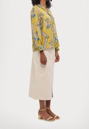 Kvinde iført en gul blomstret bluse med lange ærmer, beige midi nederdel med sidelil, og beige blokhælte sandaler, stående sidelæns.