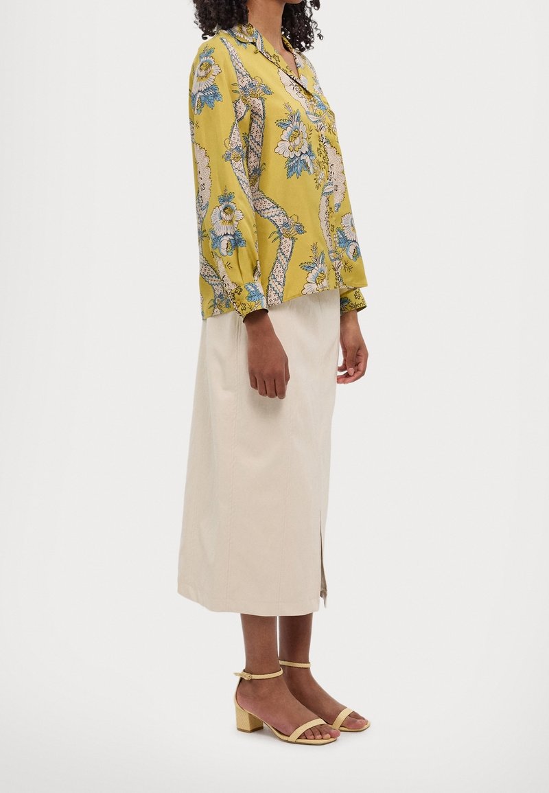 Femme portant un chemisier jaune à fleurs à manches longues, une jupe midi beige avec une fente latérale et des sandales beige à talon bloc, debout de profil.