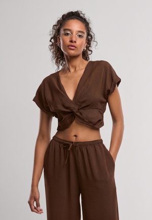 Vrouw met krullend haar die een bruin cropped top met korte mouwen en een knoop draagt en bijpassende hoge taillebroek met trekkoord, staand tegen een effen achtergrond.