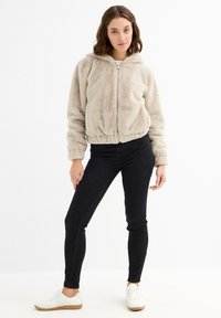 Chaqueta bomber de pelo sintético beige con capucha, cierre de cremallera en la parte frontal y cintura con elástico, combinada con jeans skinny negros y zapatillas blancas.