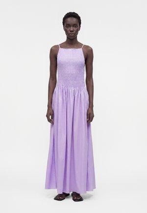 Kobieta o krótkich włosach, ubrana w bezrękawnikową, liliową, sięgającą podłogi sukienkę z marszczonym talią, stojąca na białym tle.