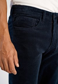 Navy kordflortbyxor med ribbad textur, med en framficka, dragkedja och bälteshällor, visade med en hand vila på sidan.