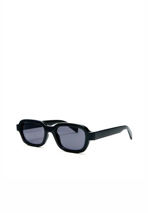 Lunettes de soleil rectangulaires noires avec des montures épaisses et des verres teintés foncés posées sur une surface blanche vues sous un angle.