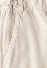 Ljus beige tyg med en slät yta, med ett elastiskt midjeband och sid sömmar för fickor. Enkelt design med minimal detaljering.