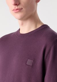 Maglione viola realizzato in un tessuto morbido, con scollatura rotonda e un piccolo logo in rilievo sul lato sinistro del petto.