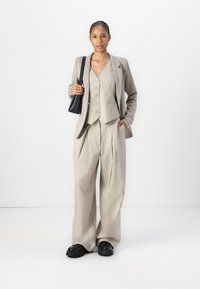 Beige taillierter Blazer über einem passenden Weste und weiten Hosen, kombiniert mit klobigen schwarzen Plateauschuhen und einer kleinen schwarzen Tasche.
