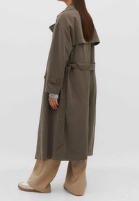 Persoon met lang bruin haar draagt een oversized grijze trenchcoat, beige broek met wijde pijpen en witte sneakers, staand tegen een effen achtergrond.