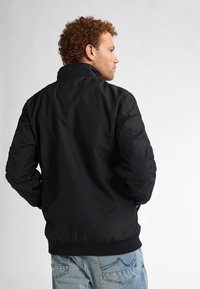 Giacca bomber nera con colletto alto, polsini e vita a coste. Texture liscia, senza motivi visibili o dettagli in metallo.