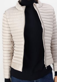 Beige pufferjakke med vandret quiltet design, høj krave og lynlås foran. Båret over en sort turtleneck og mørke jeans.