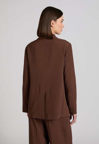 Frau mit glattem, dunkelbraunem Haar, die einen braunen Blazer mit langen Ärmeln und eine passende Hose trägt, seitlich vor einem einfarbigen Hintergrund steht.