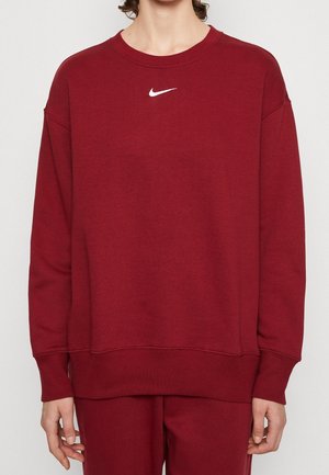 Osoba ubrana w jednolitą czerwoną bluzę Nike z małym białym logo typu swoosh na piersi, pokazana od szyi do bioder.