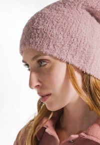 Mujer joven con piel clara y cabello rojo que lleva un gorro de punto texturizado rosa y un abrigo rosa a juego, mirando pensativamente hacia un lado.