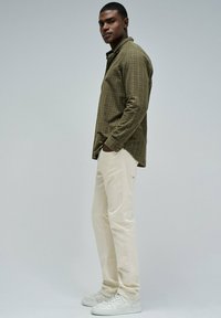 Zielona koszula w kratę, długie rękawy, z guzikami przy mankietach; kremowe spodnie, prosty krój; białe trampki; profil boczny na neutralnym tle.