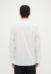 Camisa blanca de manga larga con rayas verticales marinas, con cuello clásico y puños abotonados, vista desde atrás.
