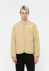 Beige polstret jakke med rund hals, frontlås og en enkelt brystlomme. Har en hvit logo på øverste høyre side.