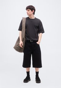 Νεαρός άνδρας που φοράει μαύρο φαρδύ μπλουζάκι και μακριές μαύρες βερμούδες, κρατώντας μια μεγάλη ώχρα-πράσινη τσάντα ώμου, στέκεται μπροστά σε λευκό φόντο.