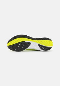 Suela de zapatilla deportiva en negro y amarillo con un agarre texturizado y multipatrón. Cuenta con una entresuela blanca y detalles de marca.