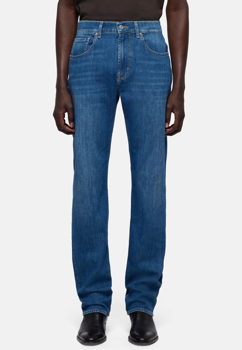 Mand iført blå jeans med lige ben, sorte støvler og en sort skjorte, stående mod en ensfarvet lys baggrund.